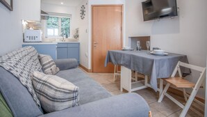 A compact living space with a sofa, a dining table with chairs, a TV mounted on the wall, and a kitchen area with cabinets and a window. - Tor Bay Cottage - 1 Bedroom - Parkmill (Swansea)