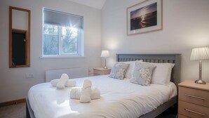 Una cama bien hecha con sábanas y toallas blancas, una mesita de noche con una lámpara, una ventana con persianas y un cuadro en la pared.