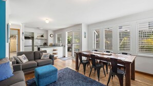 A modern living room with a sofa, ottoman, and dining table with chairs. A kitchen is visible in the background with a refrigerator and cabinets. - Price on the Beach 2c Pet-Friendly Steps to Beach (Torquay)