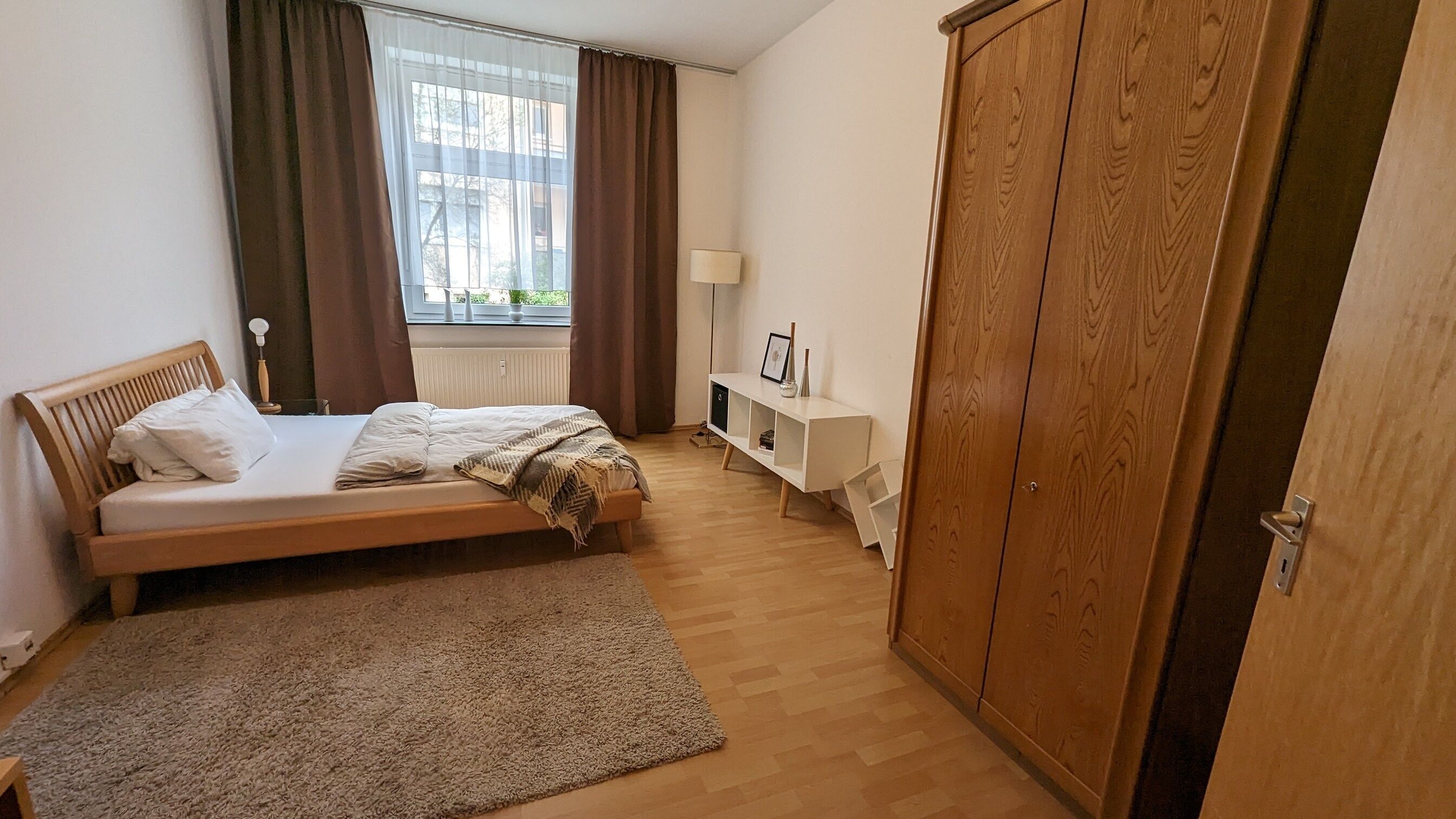 A bedroom with a wooden bed, a bedside table, a wardrobe, and a view of the outside through a window with curtains.