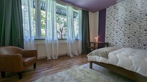Ett sovrum med trägolv, en säng, en stol, ett skrivbord och ett fönster med gardiner.