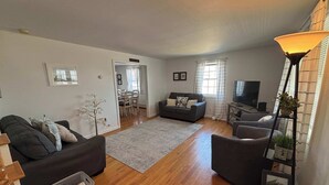 Ein Wohnzimmer mit Sofa, Sesseln und Fernseher. Im Hintergrund ist ein Essbereich zu sehen.