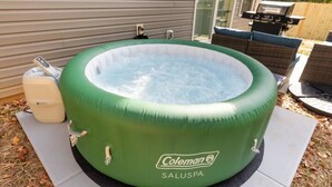Eine Coleman SALUSPA Hot Tub mit Pumpe und Bedienfeld, auf einer Terrasse mit Grill und Gartenmöbeln im Hintergrund.