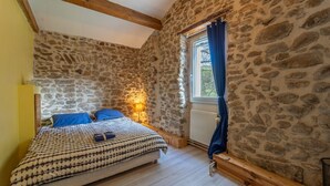 Ein Schlafzimmer mit Steinwand, einem hölzernen Bett, einem Fenster mit Vorhang und einem Nachttisch mit Lampe.