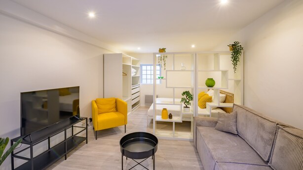 Una sala de estar moderna con un televisor de pantalla plana, un sillón amarillo, un sofá gris y una mesa de centro blanca.