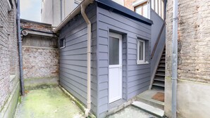Un pequeño edificio de una sola planta con fachada gris, una puerta blanca y una ventana. Se encuentra en un estrecho callejón con paredes de ladrillo a un lado y suelo de hormigón.