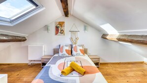 Een slaapkamer met een schuine plafond, houten balken, een dakraam, een bed met een beddegoed met patroon en een wandlamp.