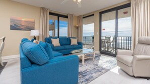 A spacious living room with a blue sofa set, a glass coffee table, and a beige armchair. A ceiling fan is visible, and the room has large windows offering a view of the ocean.