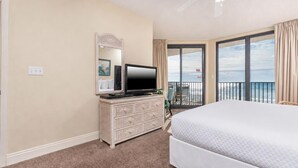 A hotel room with a bed, a television on a dresser, a balcony with a view of the ocean, and a large window.