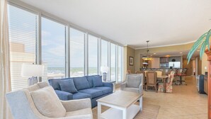 Amplia sala de estar con un sofá azul, dos sillones y una mesa de centro. Se ve un comedor con una cocina al fondo.