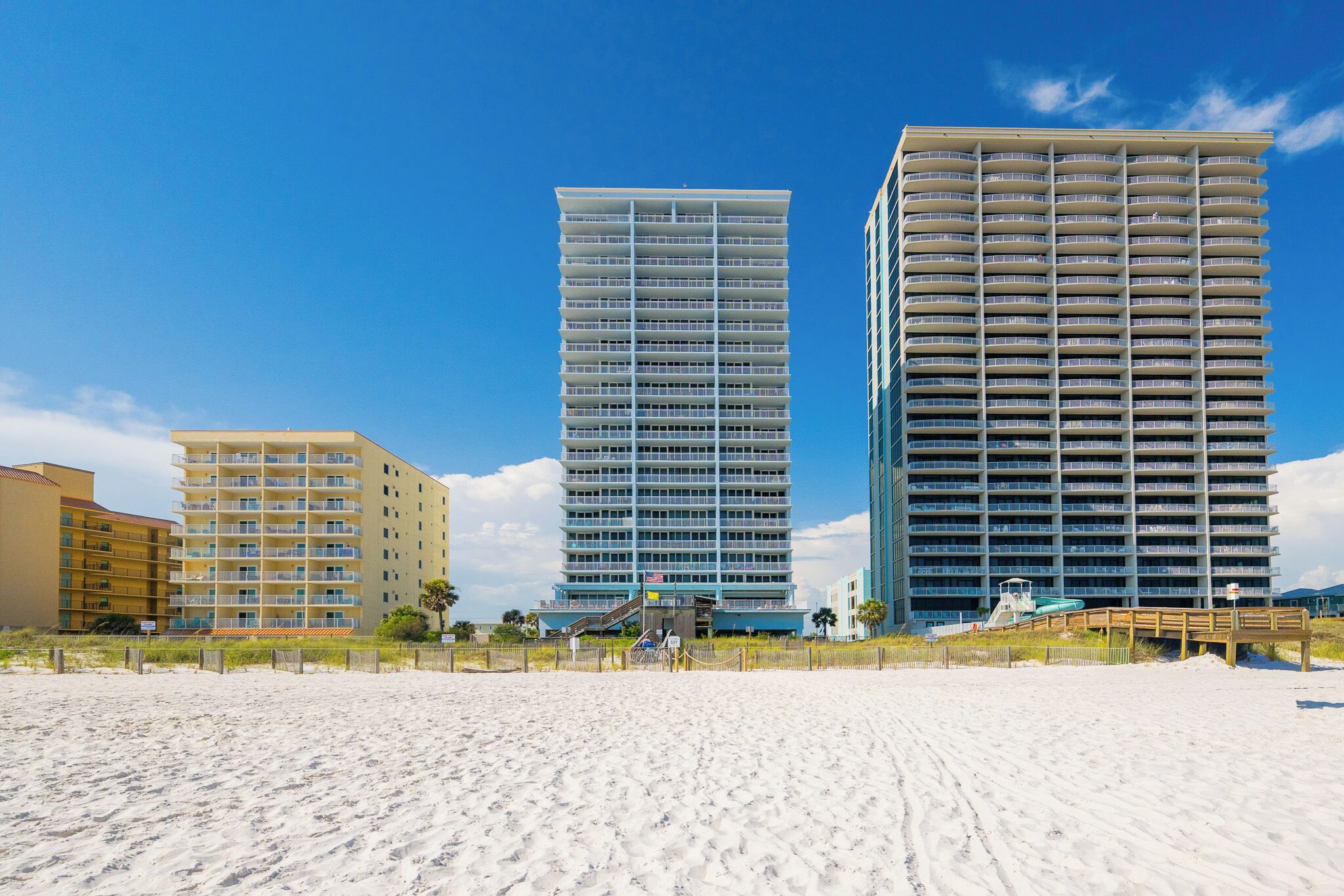 Un hotel frente a la playa con varios edificios altos, una playa de arena en primer plano y un cielo azul despejado.
