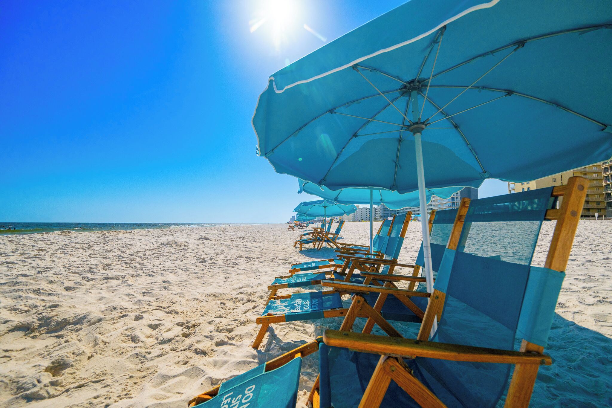Une plage bordée de rangées de chaises de plage et de parasols bleus, un ciel dégagé et une mer calme.