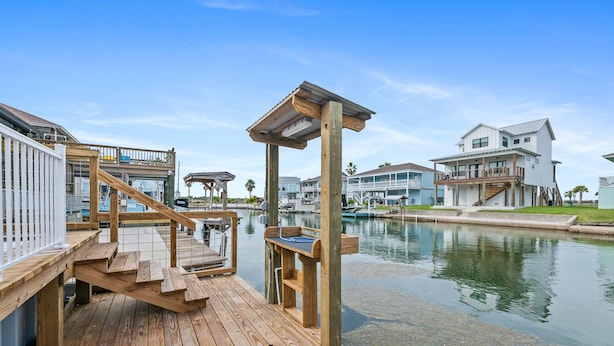 A wooden dock extending into a calm body of water, surrounded by residential buildings with balconies and a clear sky.
