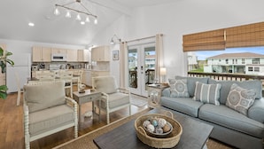 A spacious living room with a sofa, chairs, and a coffee table. There is a kitchen area with cabinets and appliances. A large window offers a view of a house.