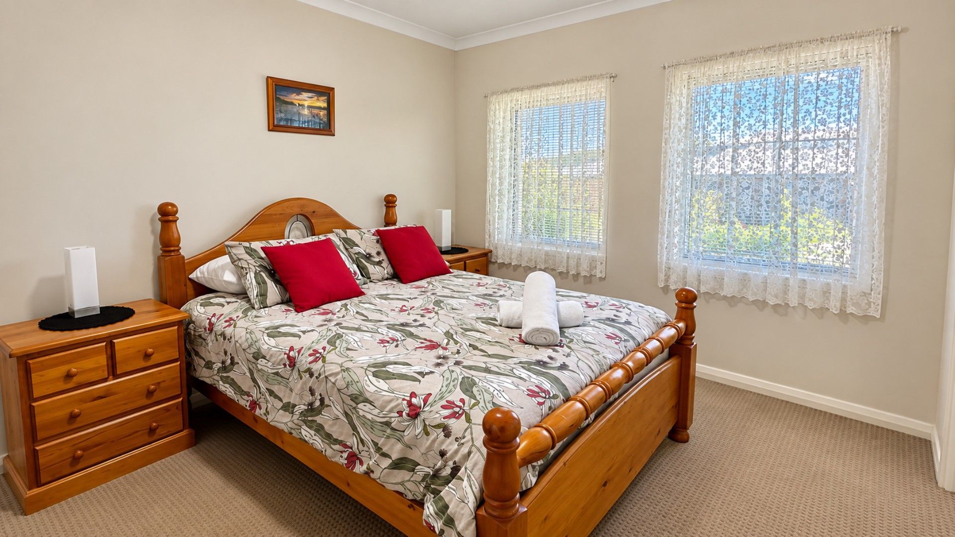 A neatly made bed with floral bedding, two bedside tables with lamps, a wooden headboard, and two windows with lace curtains.