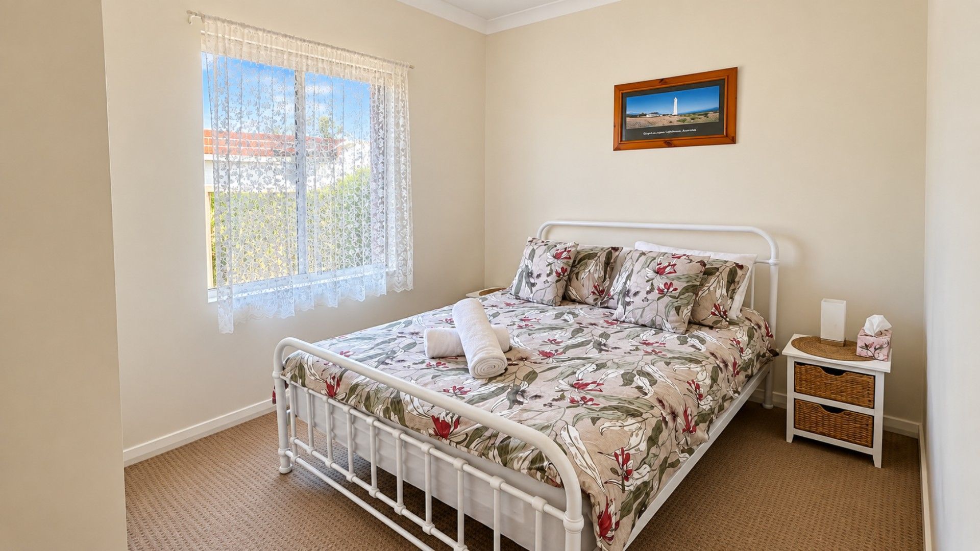 A bedroom with a metal bed frame, floral bedding, a window with lace curtains, a bedside table with a tissue box, and a framed picture on the wall.