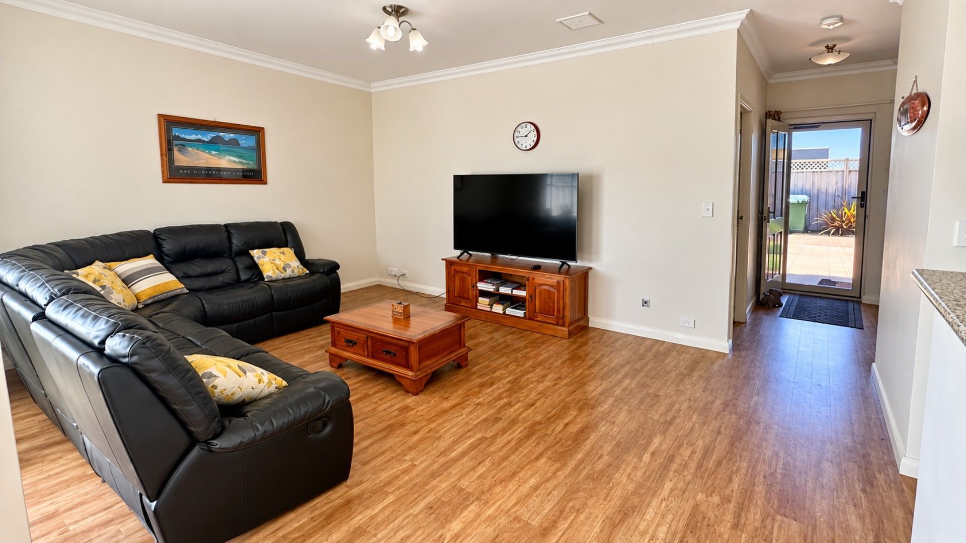 A living room with a black leather sofa, a coffee table, a flat-screen TV on a wooden stand, and a door leading to an outdoor area.