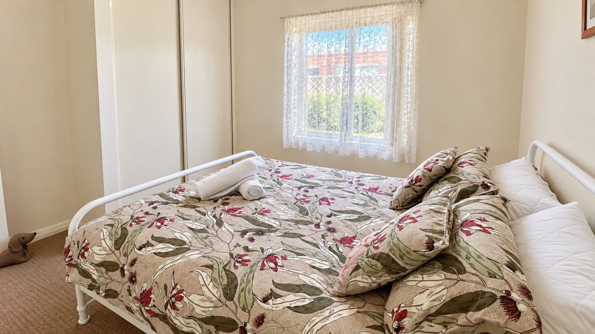 A single bed with a floral bedspread, a window with lace curtains, and a view of greenery outside.