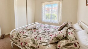 A single bed with a floral bedspread, a window with lace curtains, and a view of greenery outside.