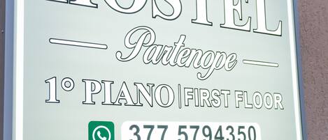 Letrero de un hostal llamado "HOSTEL Parkenop" ubicado en el primer piso, con un piano. El número de contacto es 377 5794350.