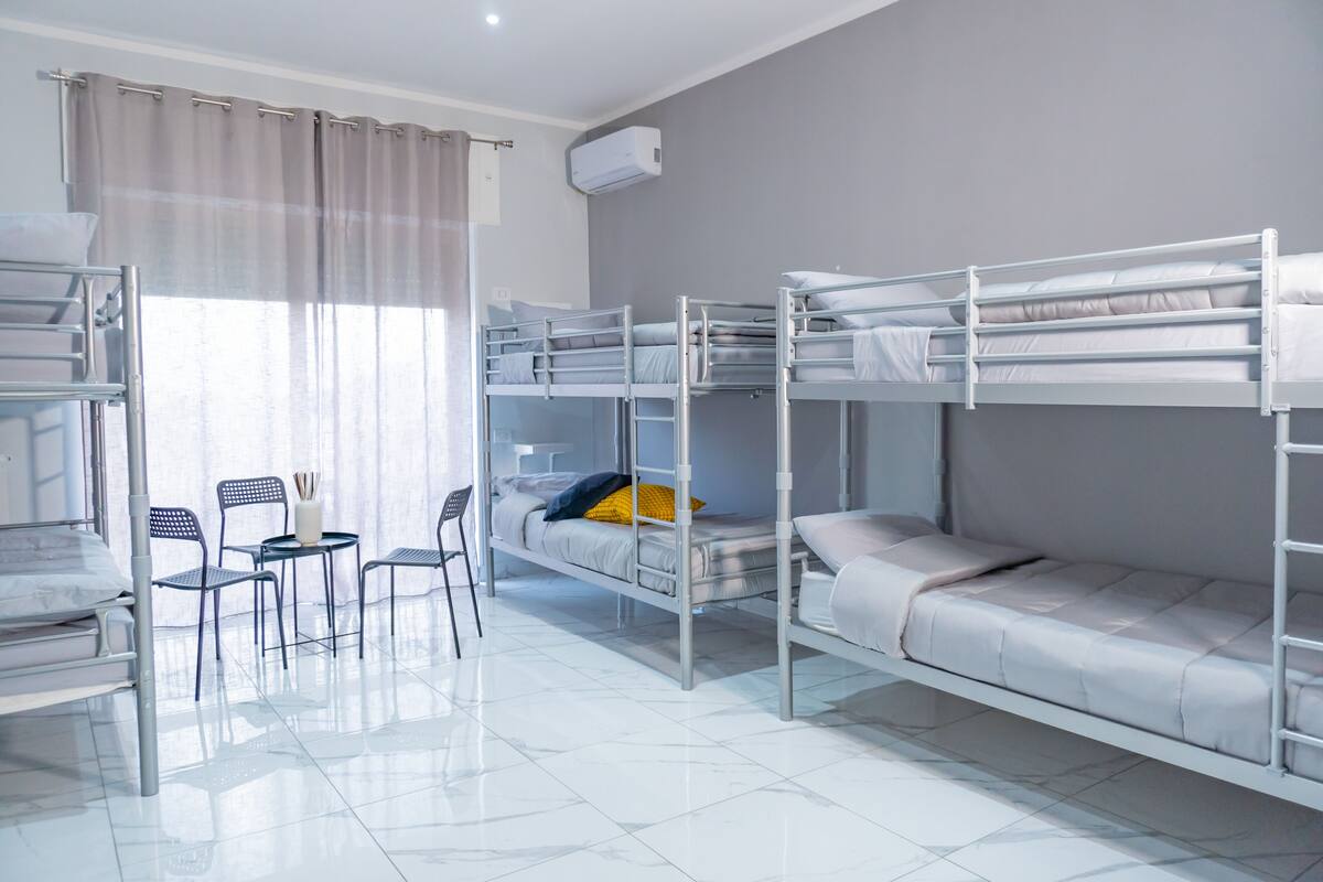 A dormitory room with bunk beds, a small table with chairs, and a window with curtains.