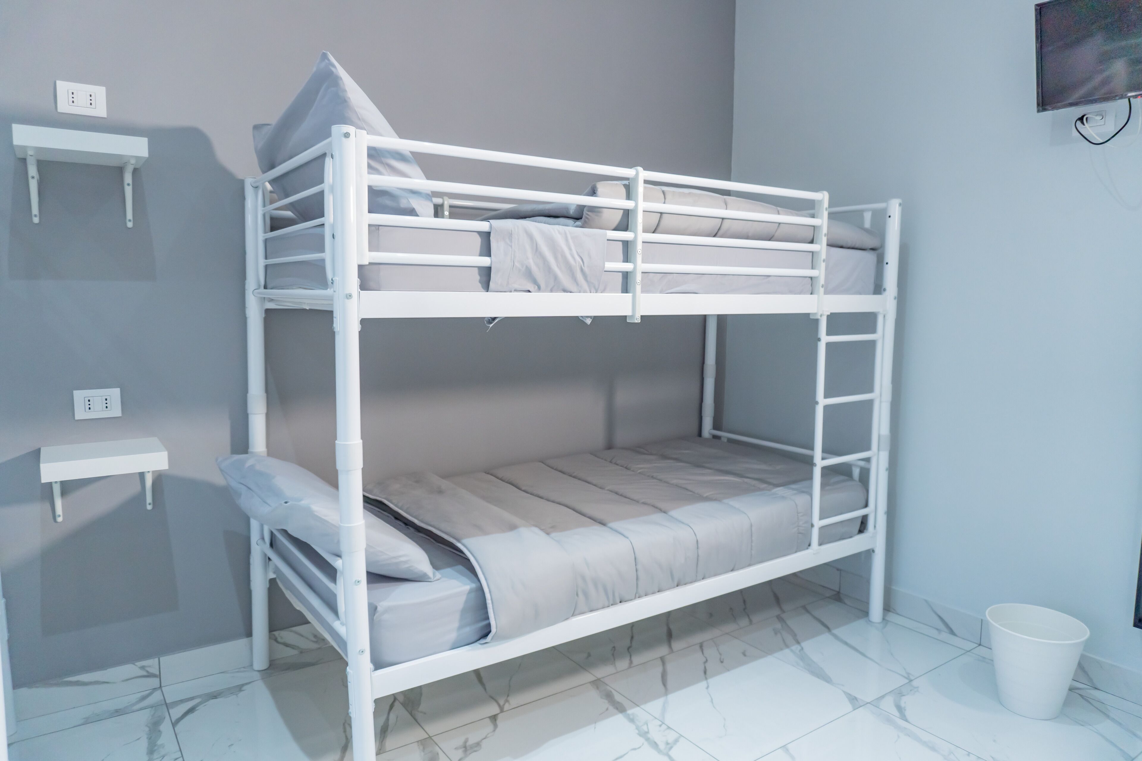 A bunk bed with a white frame, a small shelf, and a white bucket on the floor.