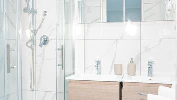 Un baño moderno con ducha envuelta en vidrio, salpicadero de mármol blanco, tocador de madera con dos lavamanos y un espejo sobre el tocador.