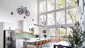 Eine moderne Küche mit einer großen Kochinsel, Barhocker und einem Essbereich mit Blick auf Bäume durch große Fenster.
