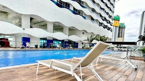 Une piscine avec des chaises longues, un bâtiment moderne à l’architecture ondulante et une vue sur le paysage urbain.