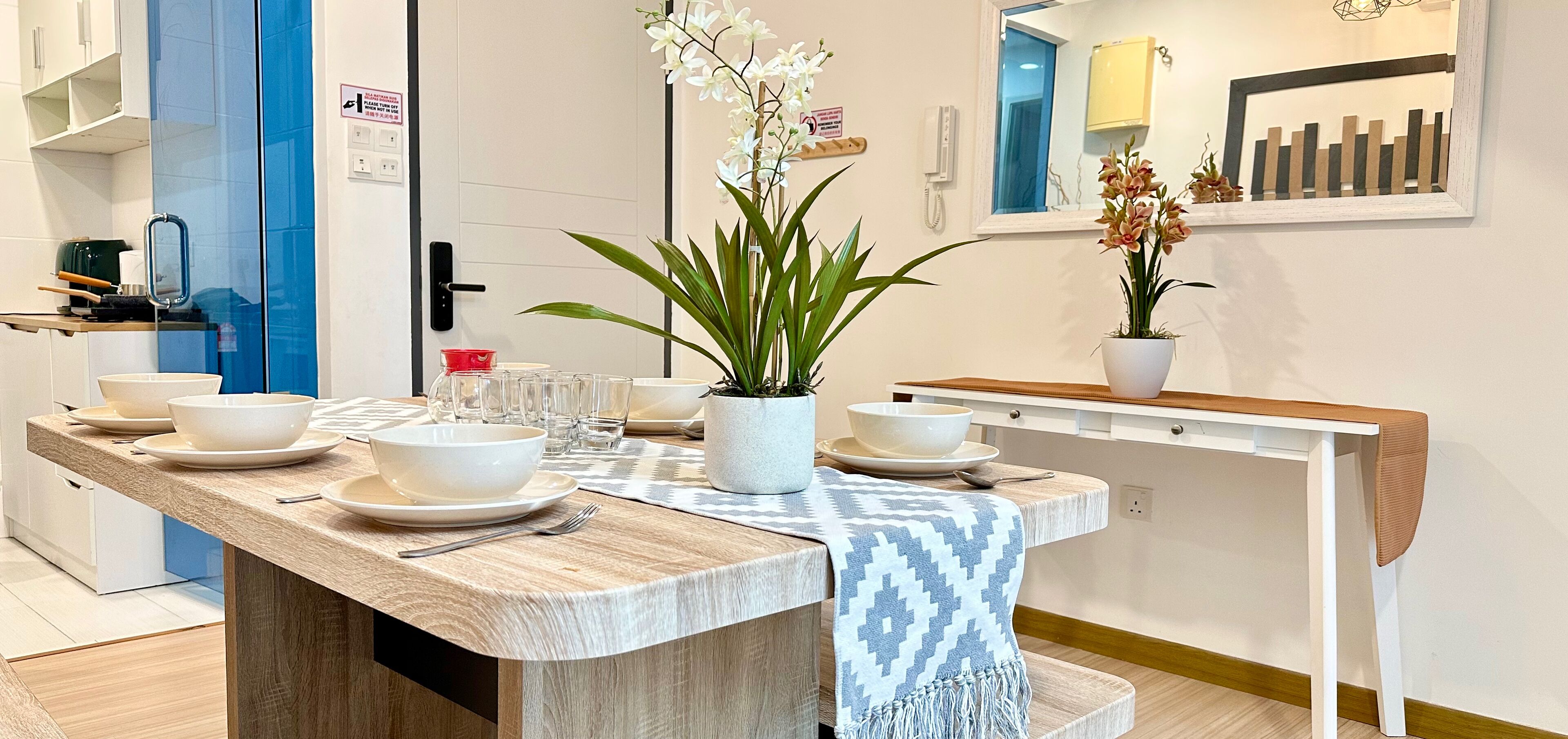 Une cuisine moderne avec un plan de travail en bois, des bols blancs et de la verrerie. On y trouve des plantes en pot et un plaid bleu et blanc posé sur la table.