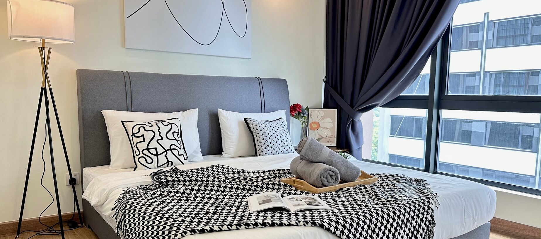 Habitación de hotel moderna con cabecera gris, manta de algodón blanco y negro, bandeja con toallas y un libro sobre la cama.