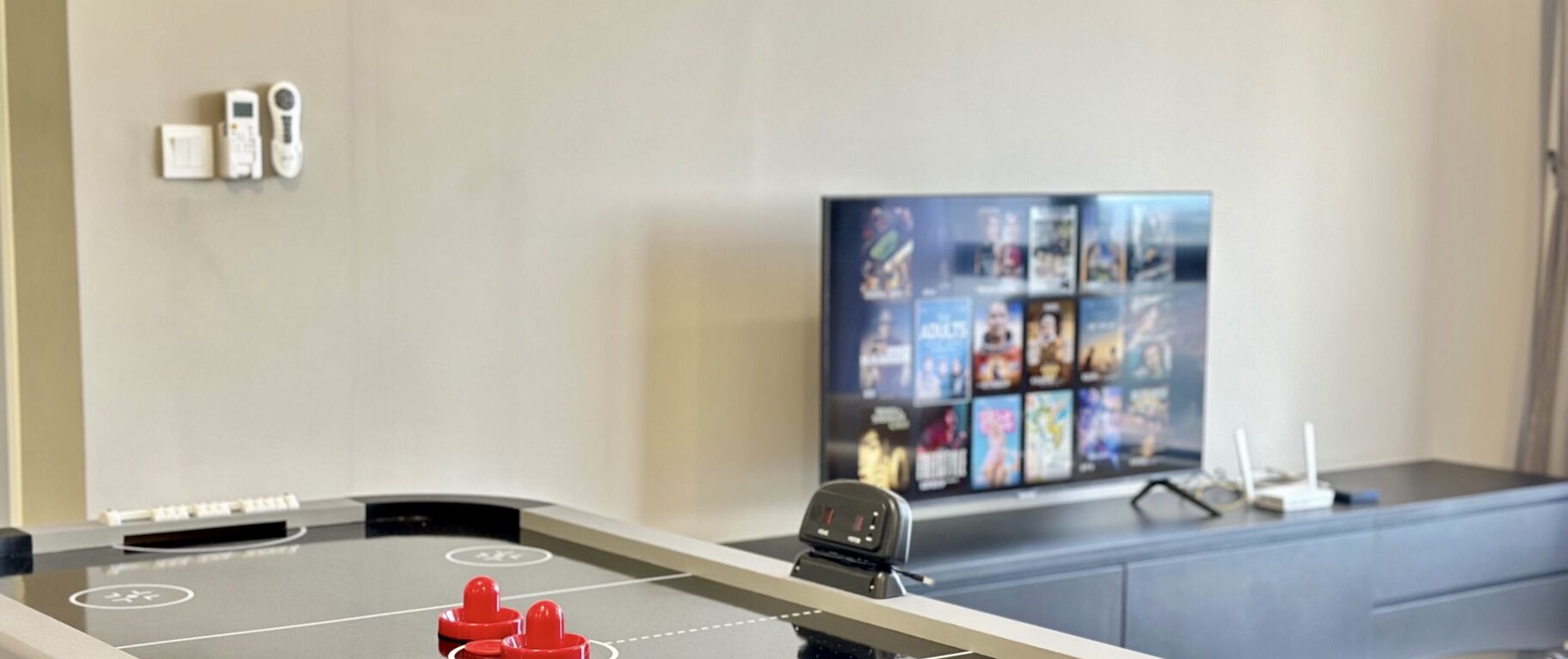 Una habitación con mesa de fútbol, televisor mostrando varios canales y escritorio con una computadora y algunos objetos encima.