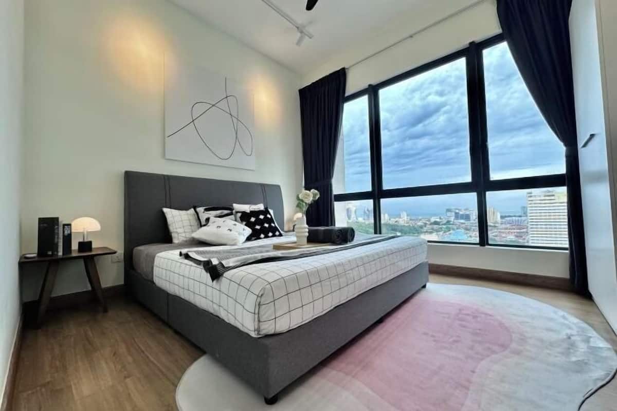 A modern bedroom with a large bed, a nightstand, a wall art piece, and a view of the city through large windows.