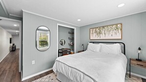 Un dormitorio con una cama grande, una mesita de noche con lámpara, un espejo en la pared y una obra de arte enmarcada sobre la cama.