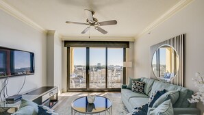 Una sala de estar moderna con un televisor de pantalla plana grande, un ventilador de techo, un sofá con cojines estampados, una mesa de centro y un ventanal con vistas a la ciudad.