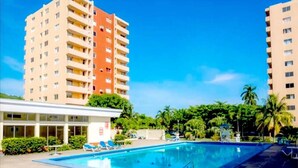 A hotel pool area with a clear blue pool, lounge chairs, and tall buildings in the background. - Beach Ready Condo than 5 mins. away from your private beach and Margaritaville. (Ocho Rios)