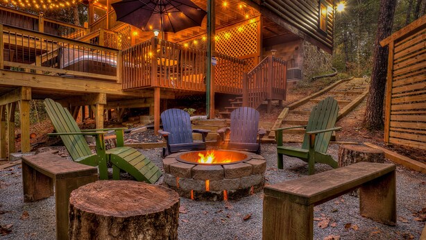 Una terraza de madera con sillas tipo Adirondack, un pozo de fuego y un área de estar cubierta con luces colgantes.