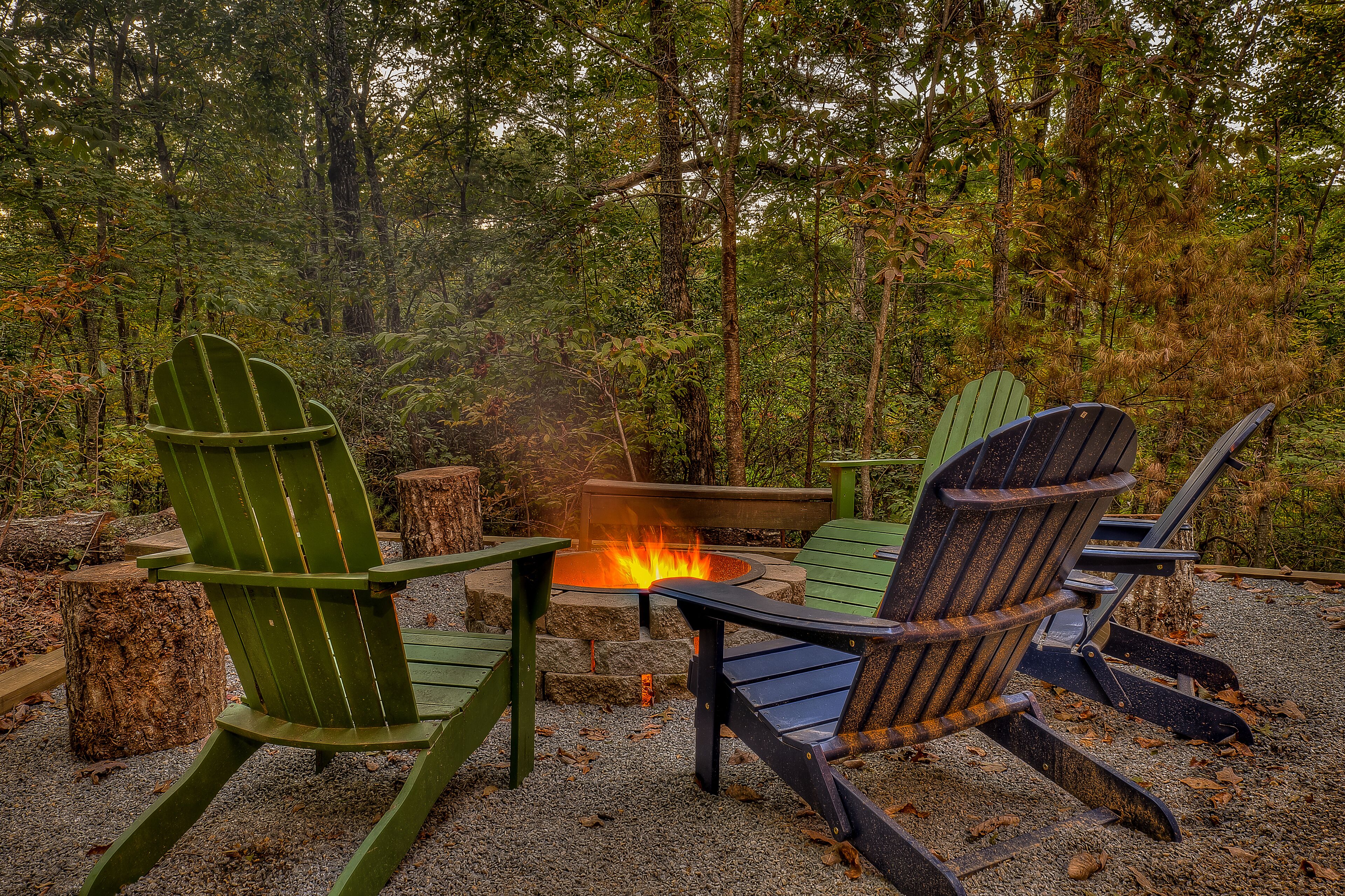 A forested area with a fire pit and Adirondack chairs.