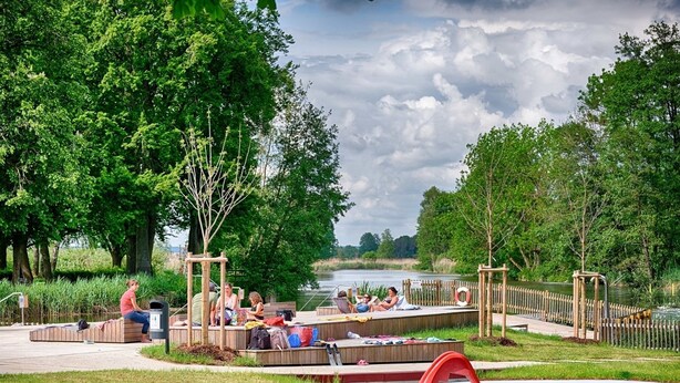 Parque a orillas del río con una pasarela de madera, una pequeña piscina y gente relajándose.