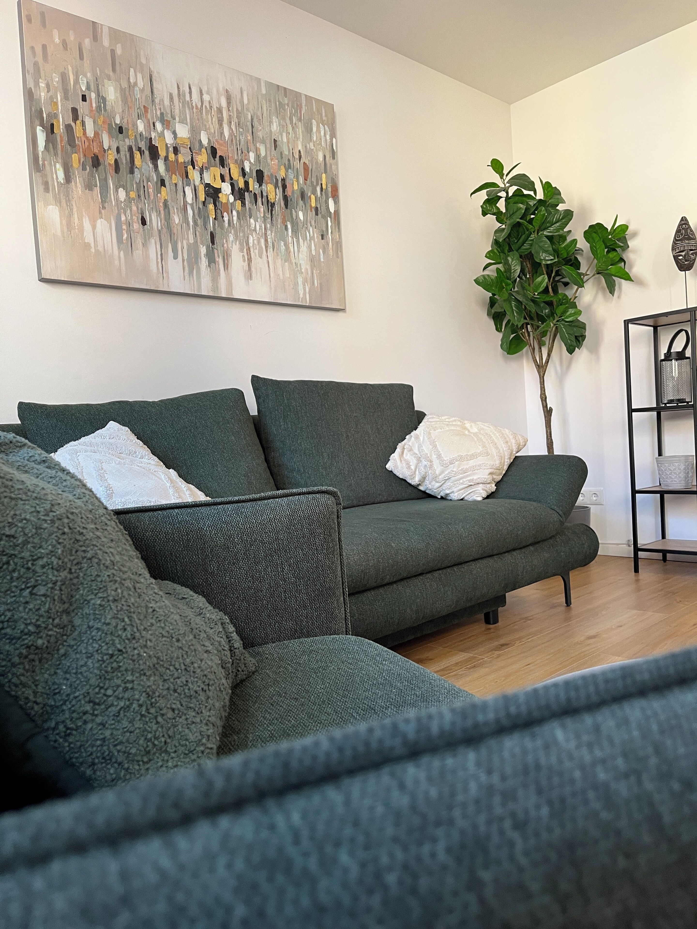 A modern living room with a green sofa, a large abstract painting, a potted plant, and a black metal shelf with decorative items.