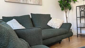A modern living room with a green sofa, a large abstract painting, a potted plant, and a black metal shelf with decorative items.