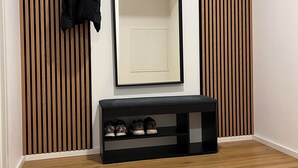 Una habitación con pared de listones de madera, un banco negro con zapatos y un espejo.