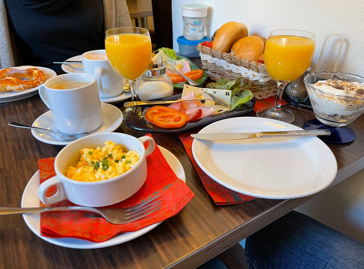 A breakfast table with plates of food, a bowl of scrambled eggs, a glass of orange juice, and a basket of bread.