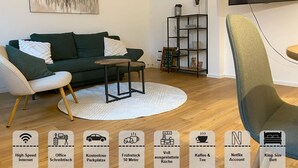 Una sala de estar moderna con un sofá verde, un sillón blanco y una mesa de centro redonda. Un televisor de pantalla plana en la pared muestra 
