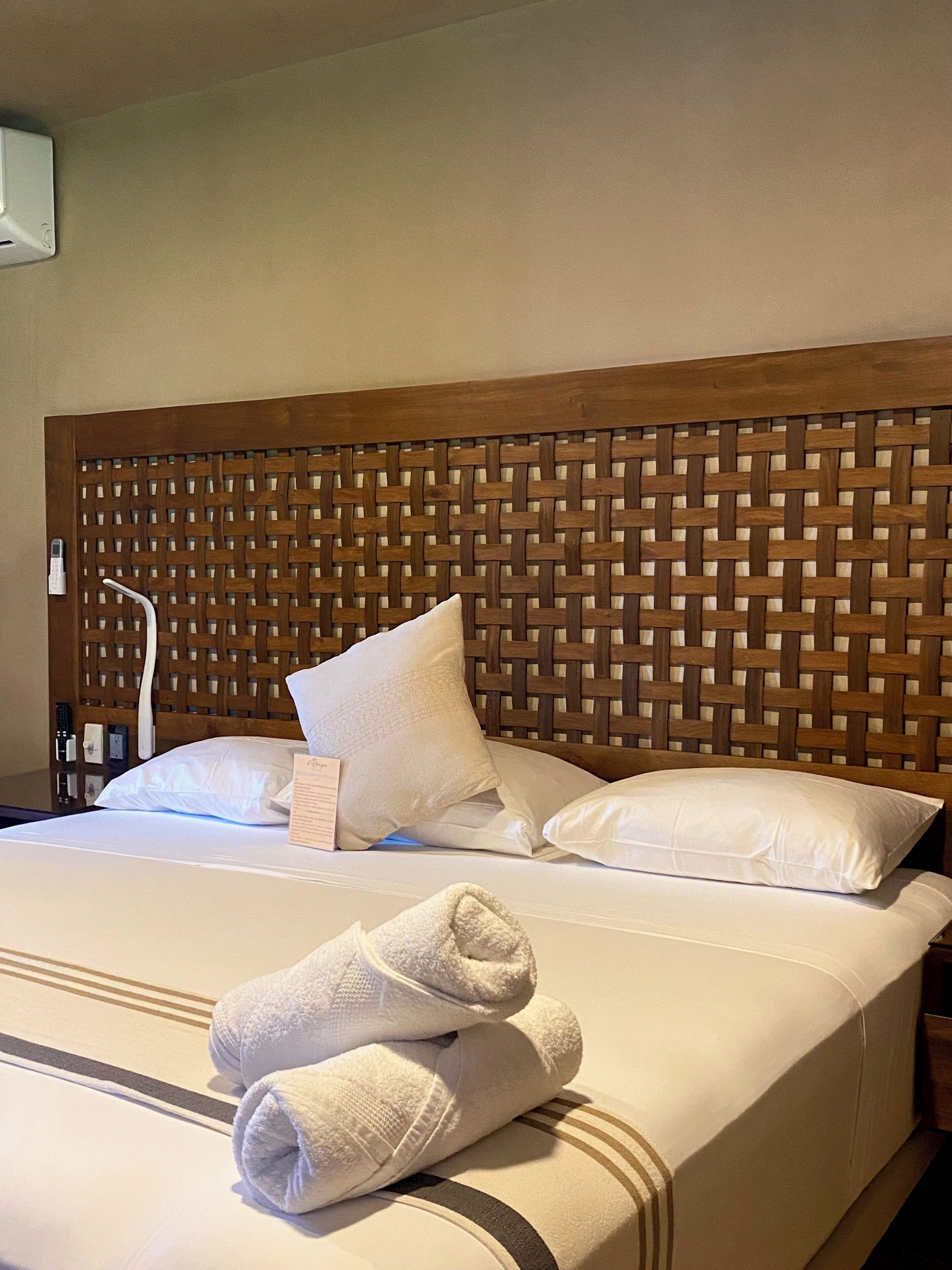 A hotel room with a wooden headboard, white bedding, and a neatly folded towel on the bed.