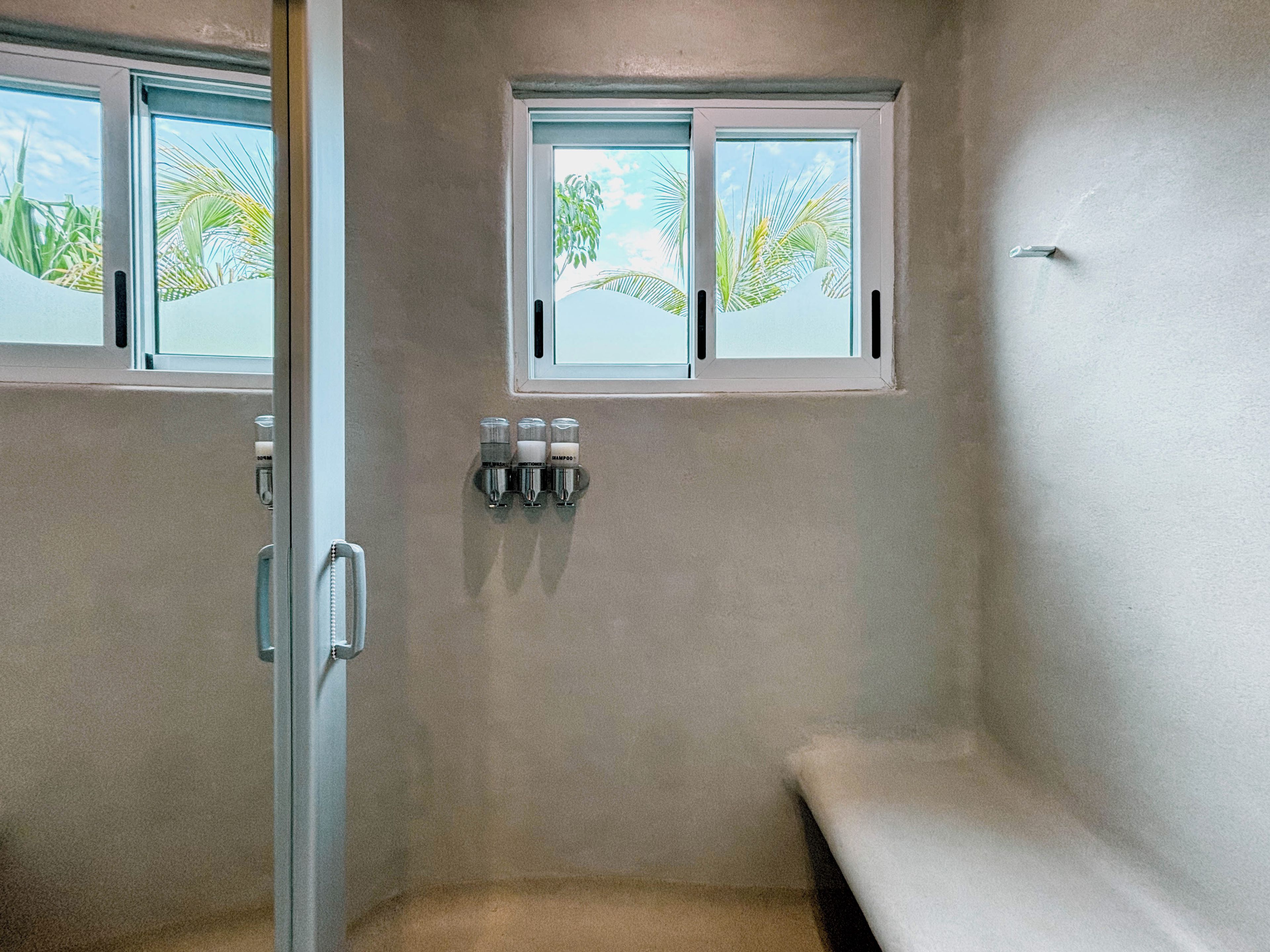 A bathroom with a shower, a bench, and two windows with a view of palm trees.