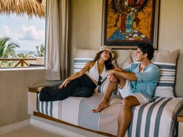 Una pareja sentada en una cama con ropa de cama a rayas, un cuadro en la pared y vistas al exterior.