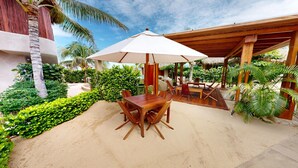 A wooden deck with a table and chairs under a white umbrella, surrounded by lush greenery and a thatched-roof structure. - El Origen Villas & Spa Puerto Escondido (San Pedro Mixtepec)