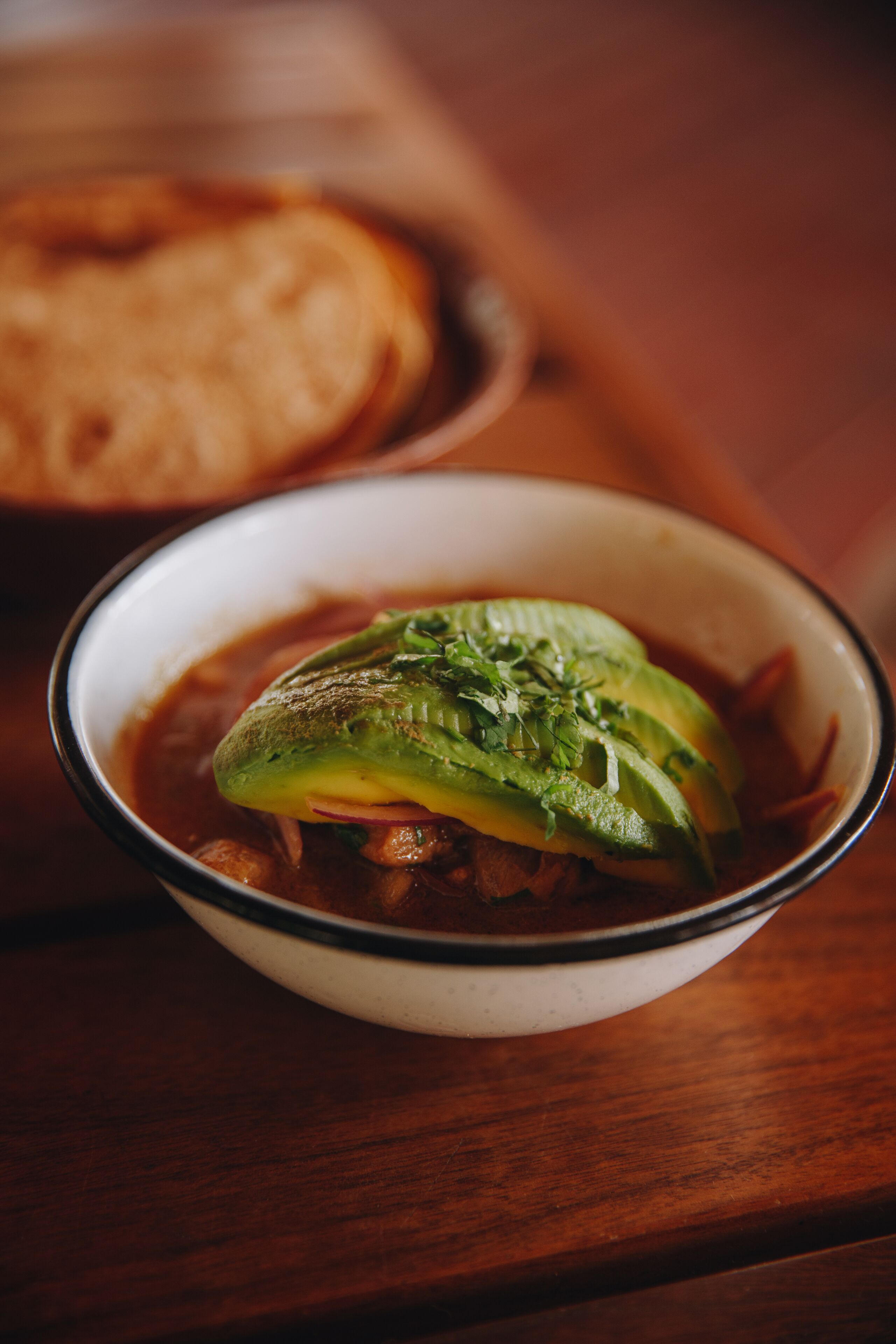 A bowl of soup with a green topping, possibly avocado, on a wooden table.