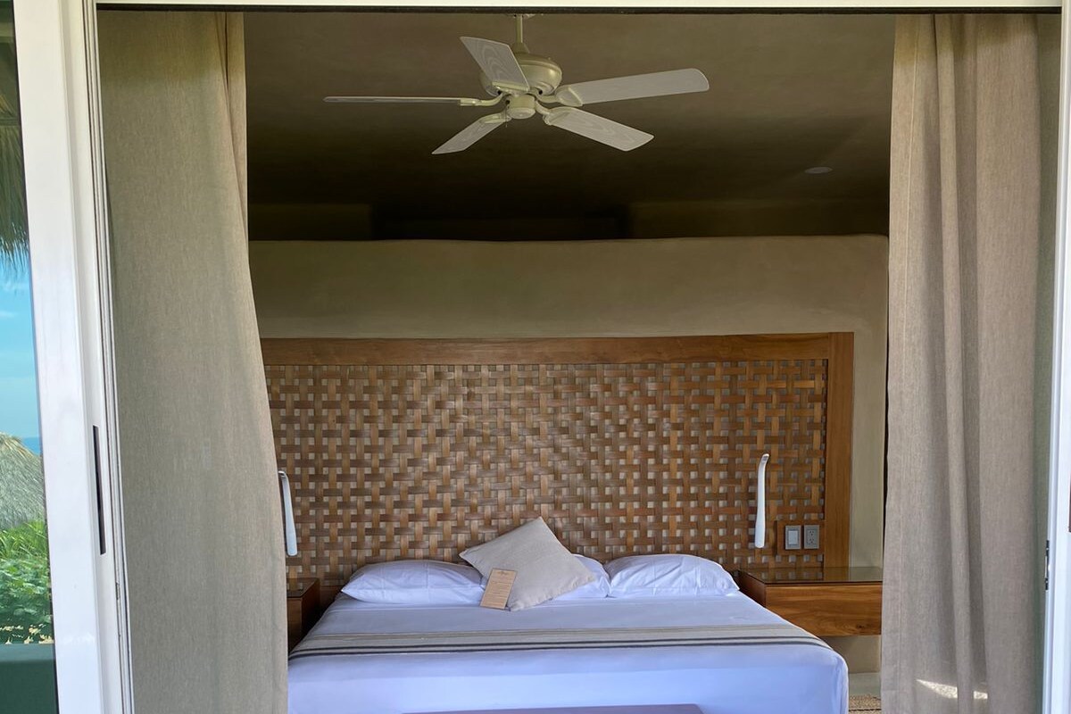 A bedroom with a bed, a wooden bench, a ceiling fan, and a view through a window.